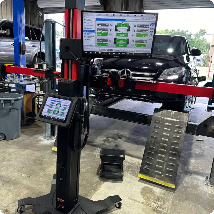 A black car is elevated on a vehicle lift in a garage. In the foreground, a computerized wheel alignment machine with two screens displays diagnostic information. Various tools and equipment are visible around the workspace.