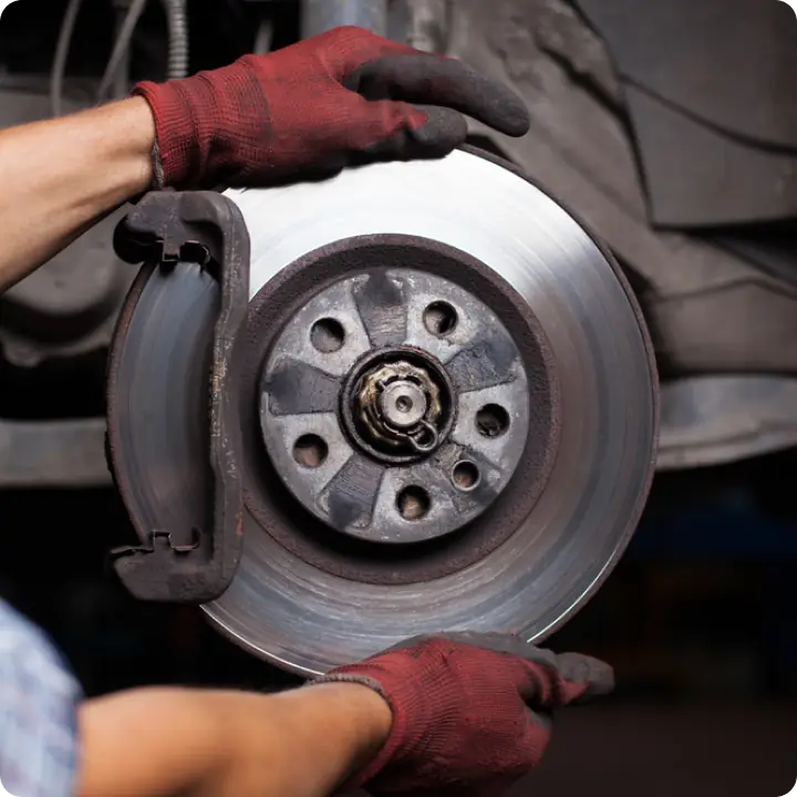 A person wearing red gloves is installing or removing a car brake disc and caliper, with the wheel removed, exposing the vehicle’s braking system.