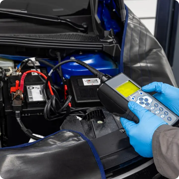 A person wearing blue gloves uses a diagnostic scanner to check the battery of a car with its hood open in an automotive workshop.