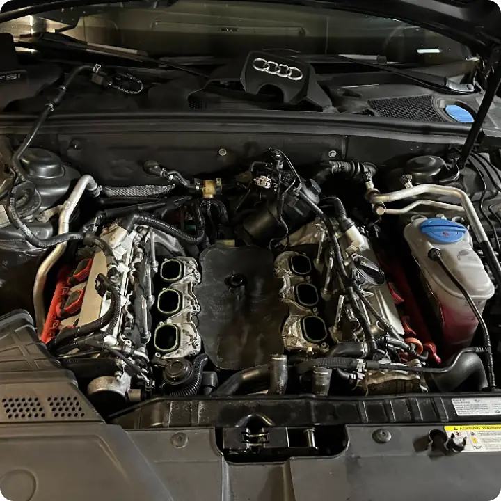 The engine bay of an Audi vehicle with the intake manifold removed, revealing cylinder banks and various engine components, hoses, and wiring. The Audi logo is visible on the engine cover at the back.