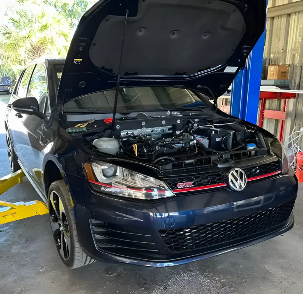 A dark blue Volkswagen GTI in My Car Guy of Plant City, with its hood open, is parked in a garage, elevated on a car lift, exposing the engine for maintenance or repair.