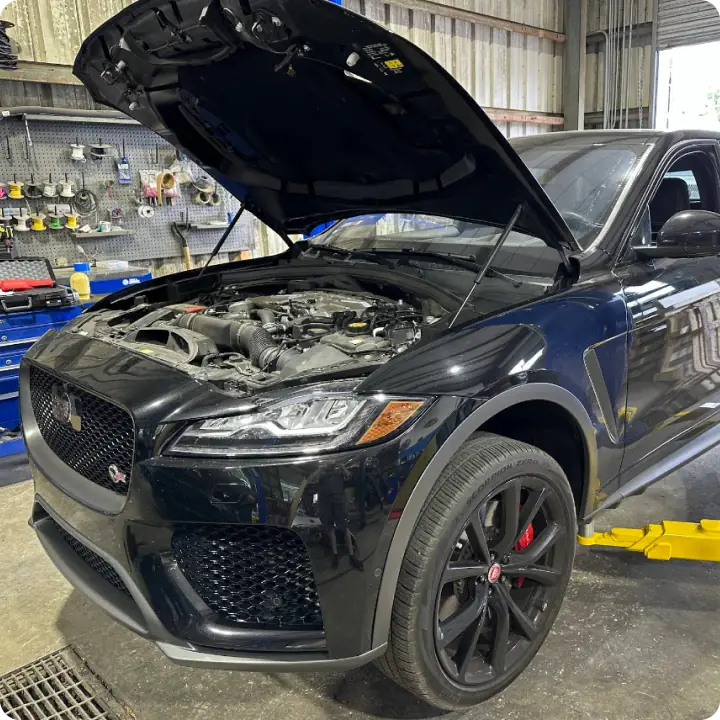 A black Jaguar SUV with its hood open is parked inside an auto repair shop. The car is on a hydraulic lift, and tools and equipment are visible on the wall in the background.