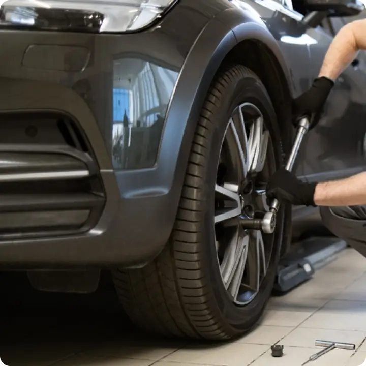 A person uses a wrench to tighten the lug nuts on a cars front wheel in a garage. The car is lifted with a jack, and tools are scattered on the tiled floor nearby.