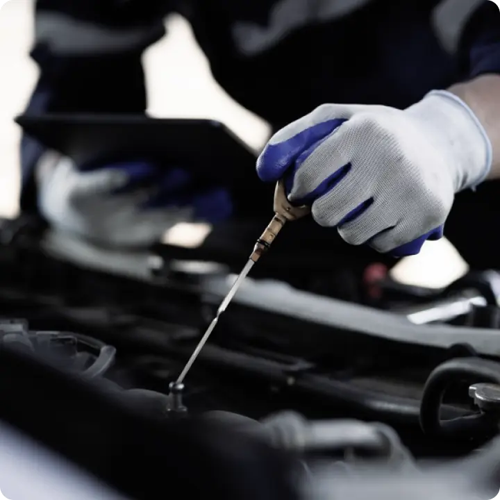 A person wearing white and blue gloves checks an engines oil level with a dipstick while holding a tablet above an open car hood.