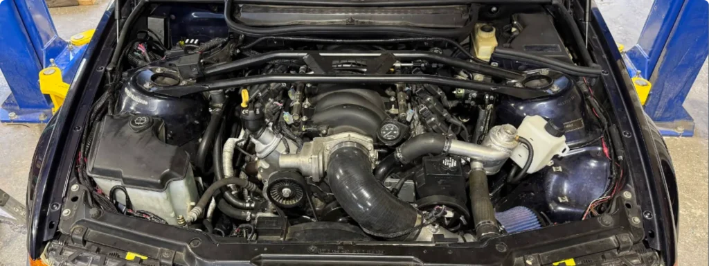 A car engine bay with a visible turbocharger setup, various hoses, and components. The car is on a lift, with blue lift arms visible on both sides. The engine appears clean and is centrally positioned.