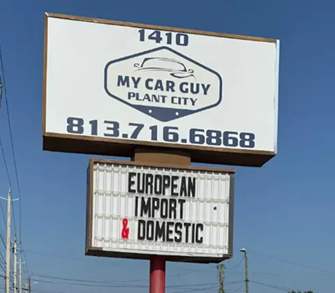 A roadside sign for My Car Guy Plant City displays their phone number and advertises services for European, import, and domestic vehicles against a clear blue sky.