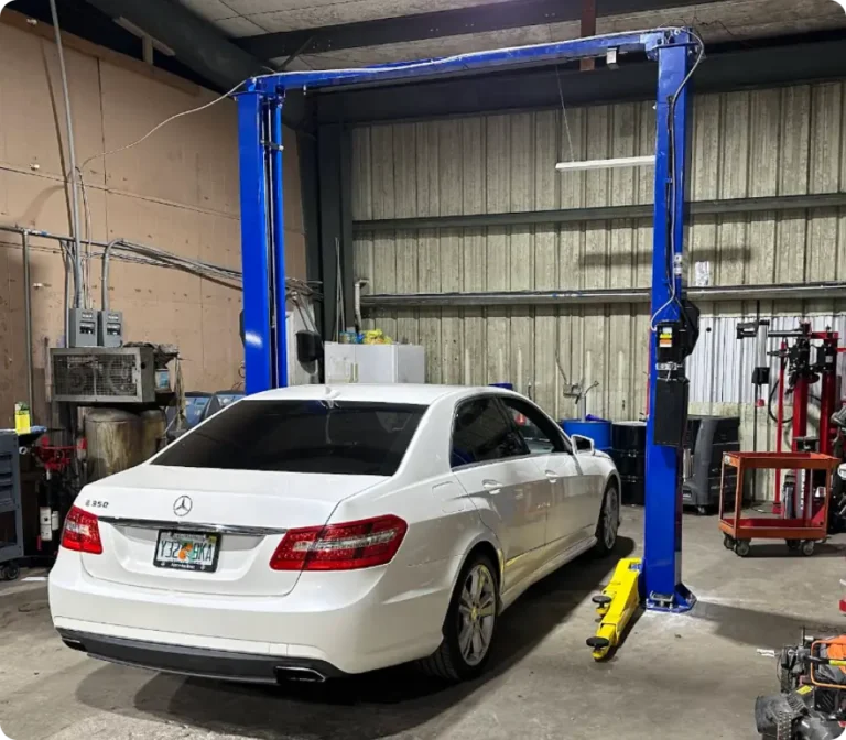 A white Mercedes-Benz E350 parked inside an auto repair shop under a blue car lift, surrounded by tools and equipment in My Car Guy of Plant City.