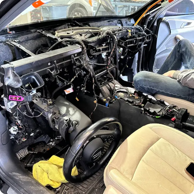 A car interior with the dashboard completely removed, exposing wires and internal components. A person sits in the passenger seat working on the disassembled dashboard area. A yellow cloth is on the floor.