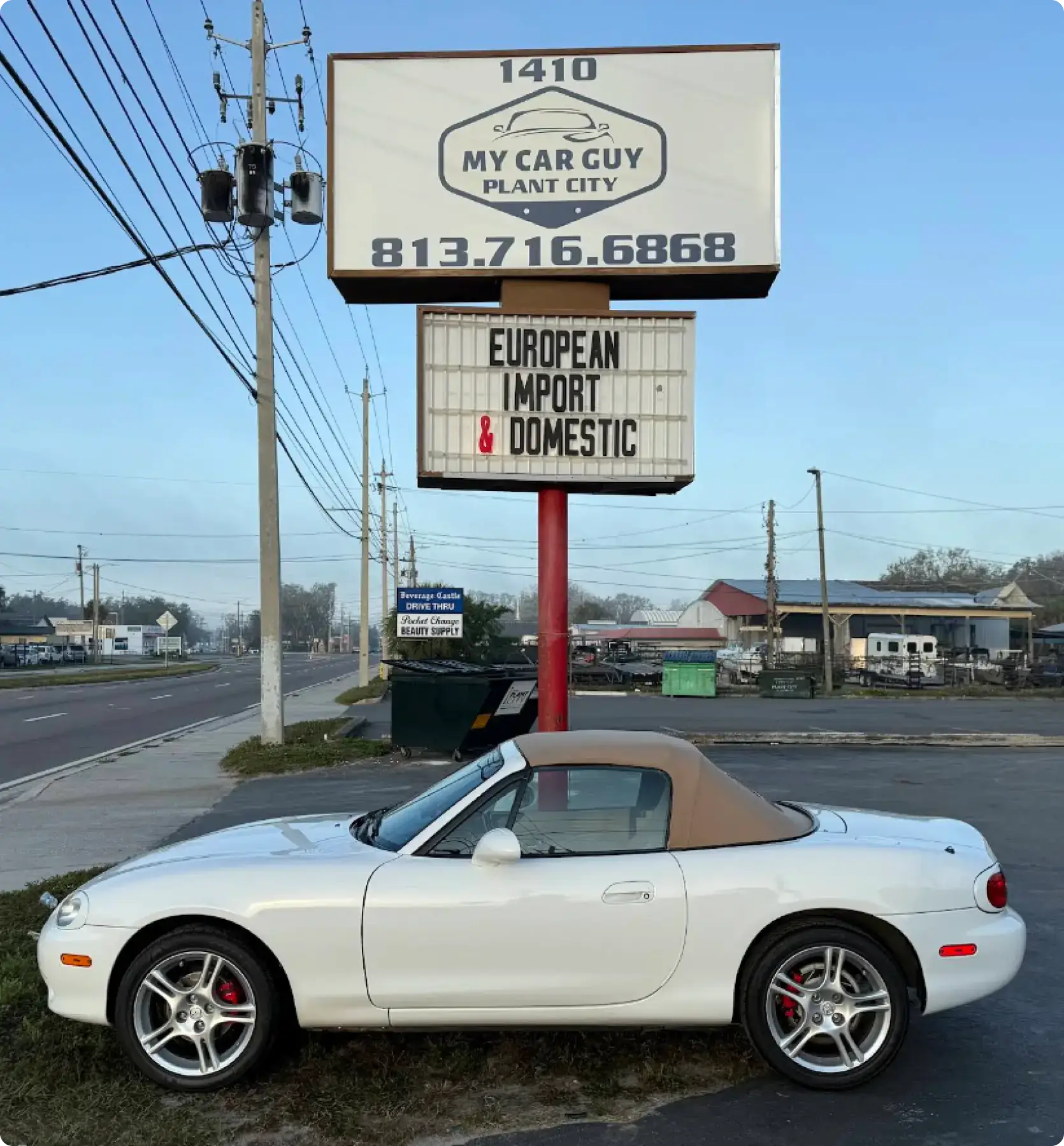 A white convertible sports car is parked in front of a car dealership sign that reads MY CAR GUY PLANT CITY and EUROPEAN IMPORT & DOMESTIC, with a phone number and street in the background.