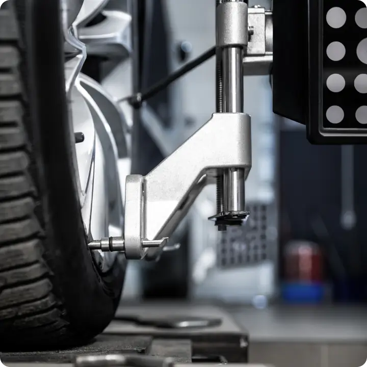 Close-up of a car wheel being aligned with a precision alignment tool clamped to the tire in an automotive workshop. The tool and tire are in focus, with a blurred background of garage equipment.