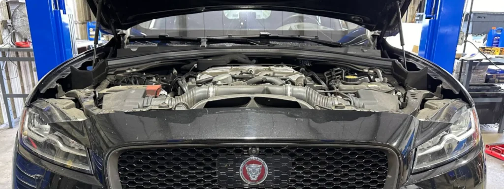 A black Jaguar car with its hood open, exposing the engine bay, is parked inside a garage with various tools and equipment visible in the background in My Car Guy auto repair Plant City FL.