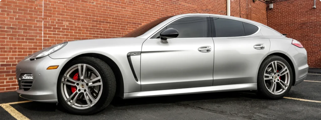 A silver Porsche Panamera sedan is parked in an outdoor lot next to a brick wall, with tinted windows and red brake calipers visible.
