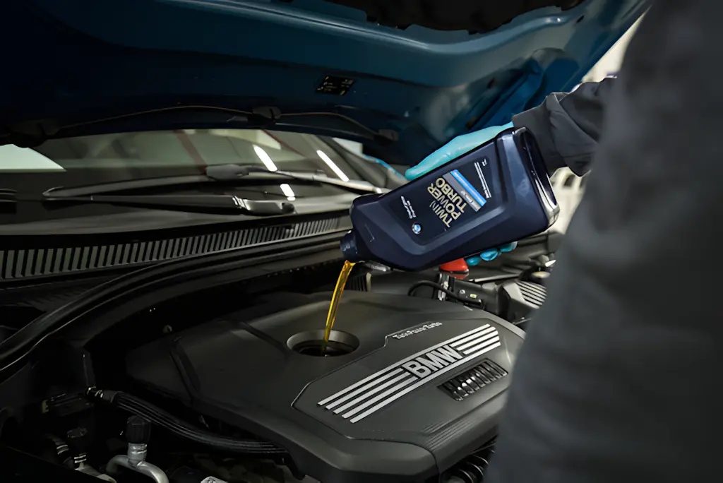 A person wearing gloves pours engine oil from a blue bottle labeled "Twin Power Turbo" into the engine of a BMW car with the hood open.
