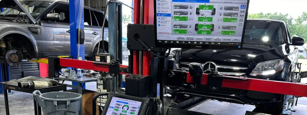 A car repair shop with a black car undergoing a wheel alignment, monitors displaying alignment data, and a silver SUV raised on a hydraulic lift in the background. Various tools and equipment are visible around the shop.