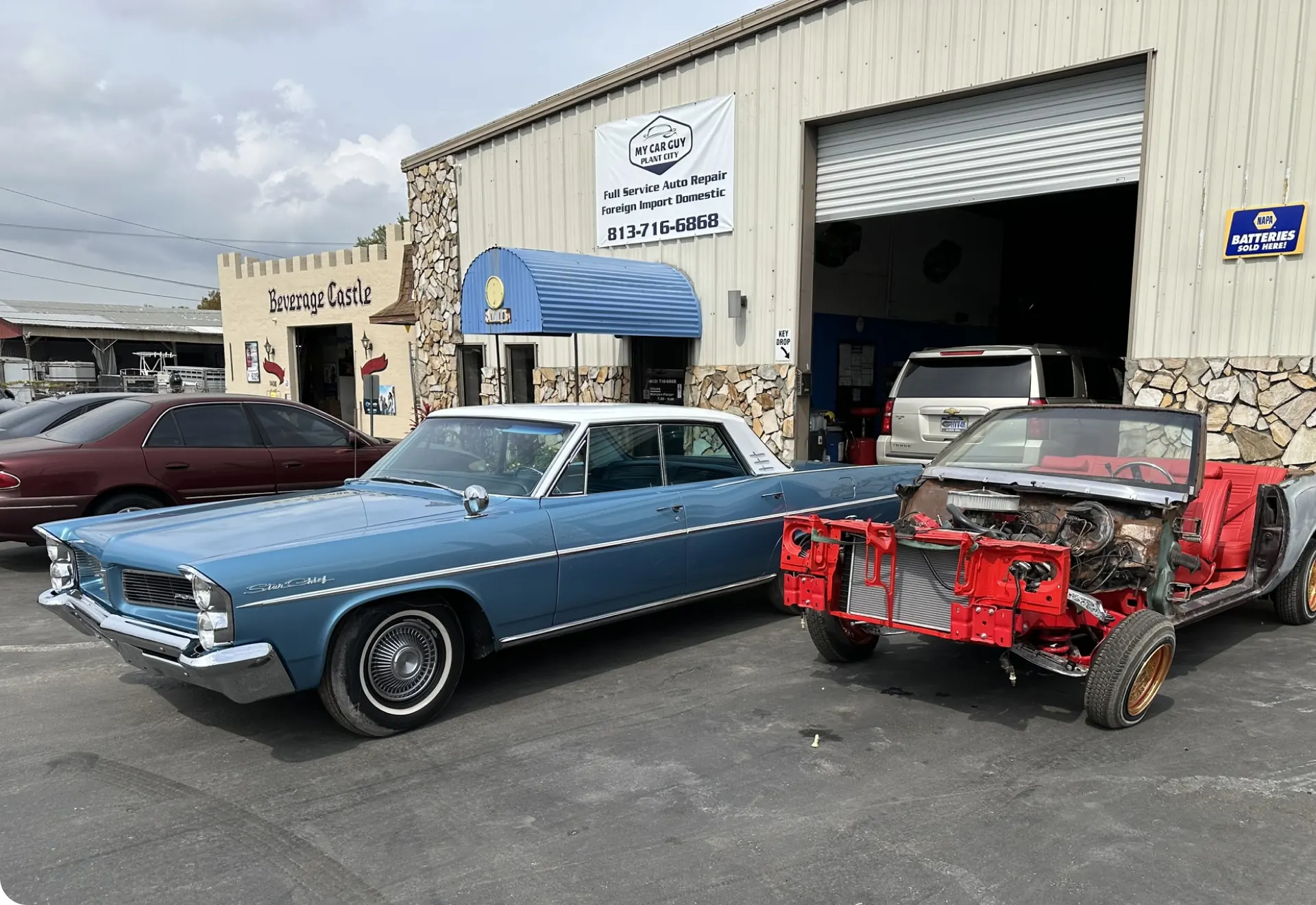 A blue vintage car and a stripped red car frame are parked outside an auto repair shop with an open garage door and a sign displaying the shop’s name and phone number. Other vehicles are visible in the background.