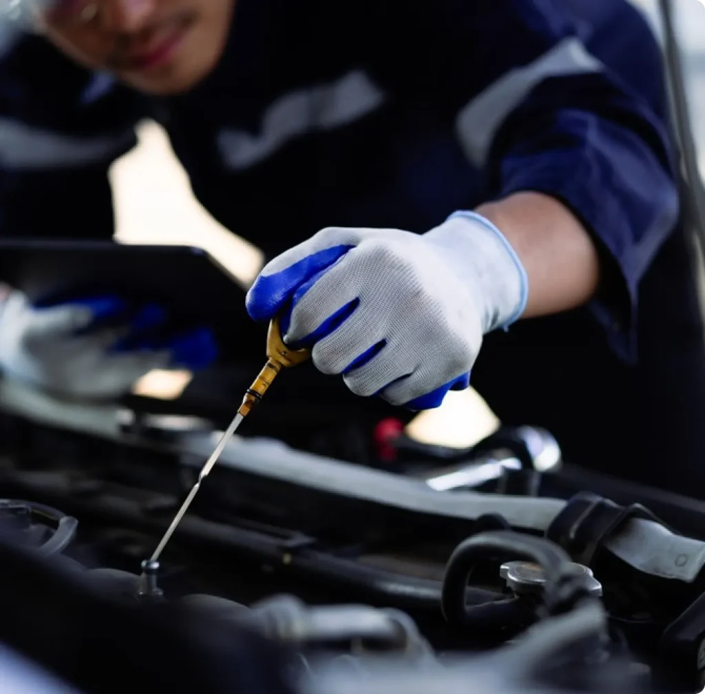 A person wearing gloves checks a car’s oil level with a dipstick, while holding a tablet in the other hand, standing over the open engine bay.