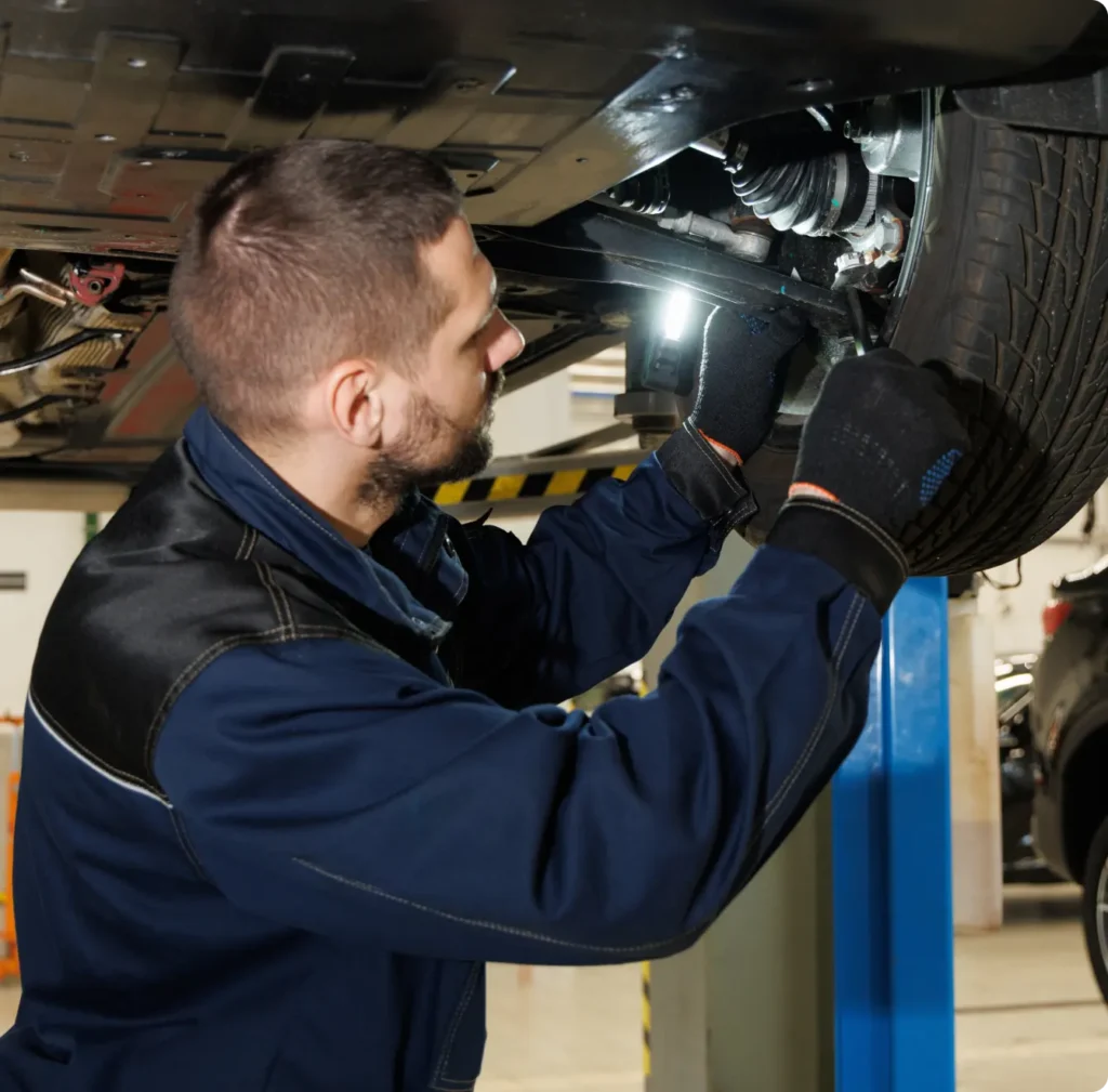 A mechanic in a blue uniform and gloves inspects the underside of a car on a lift, using a flashlight to examine parts near the wheel.