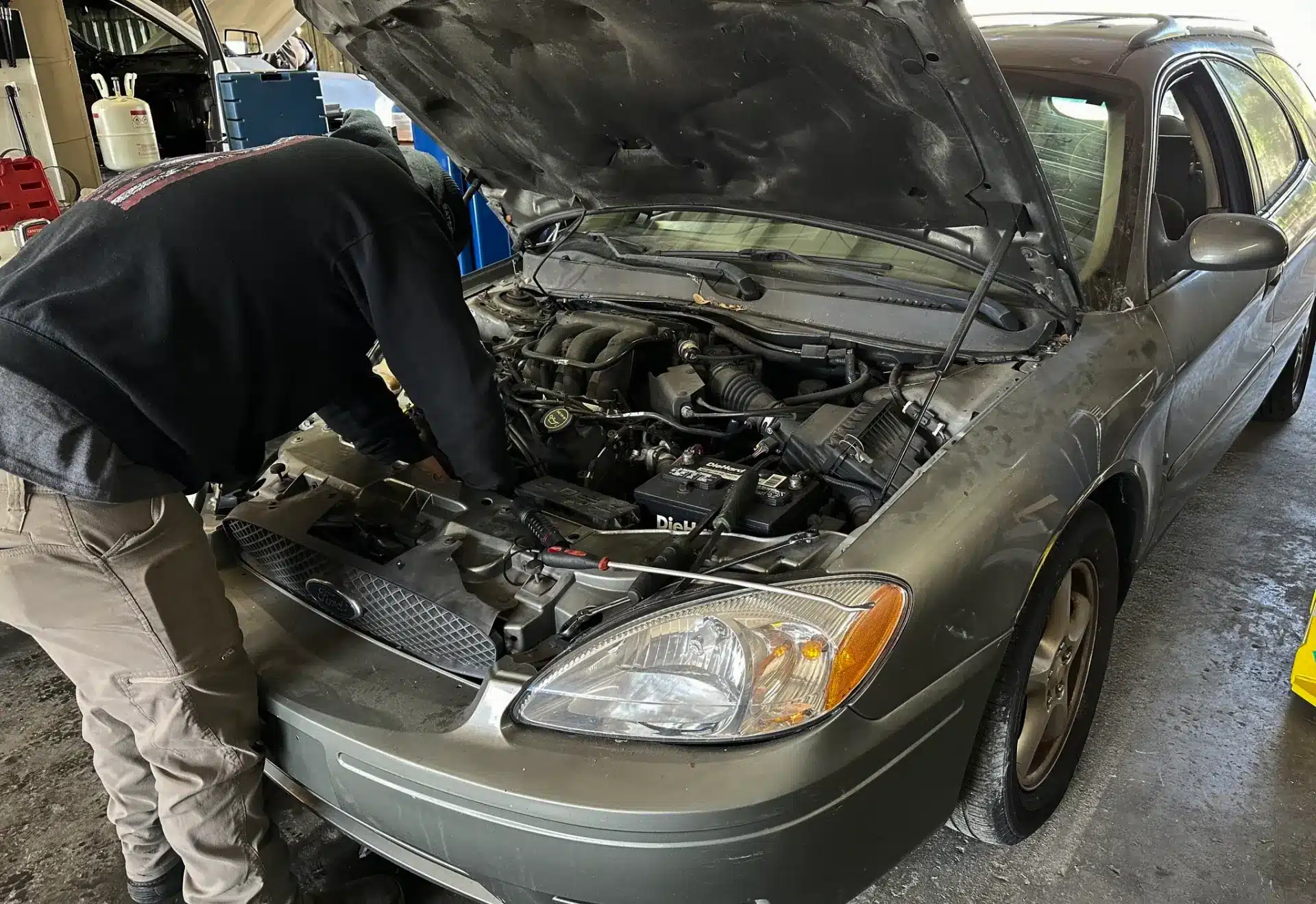 A person works under the hood of a gray sedan in a garage, performing maintenance or repairs on the engine. The cars hood is open and various tools and equipment are visible in the background.