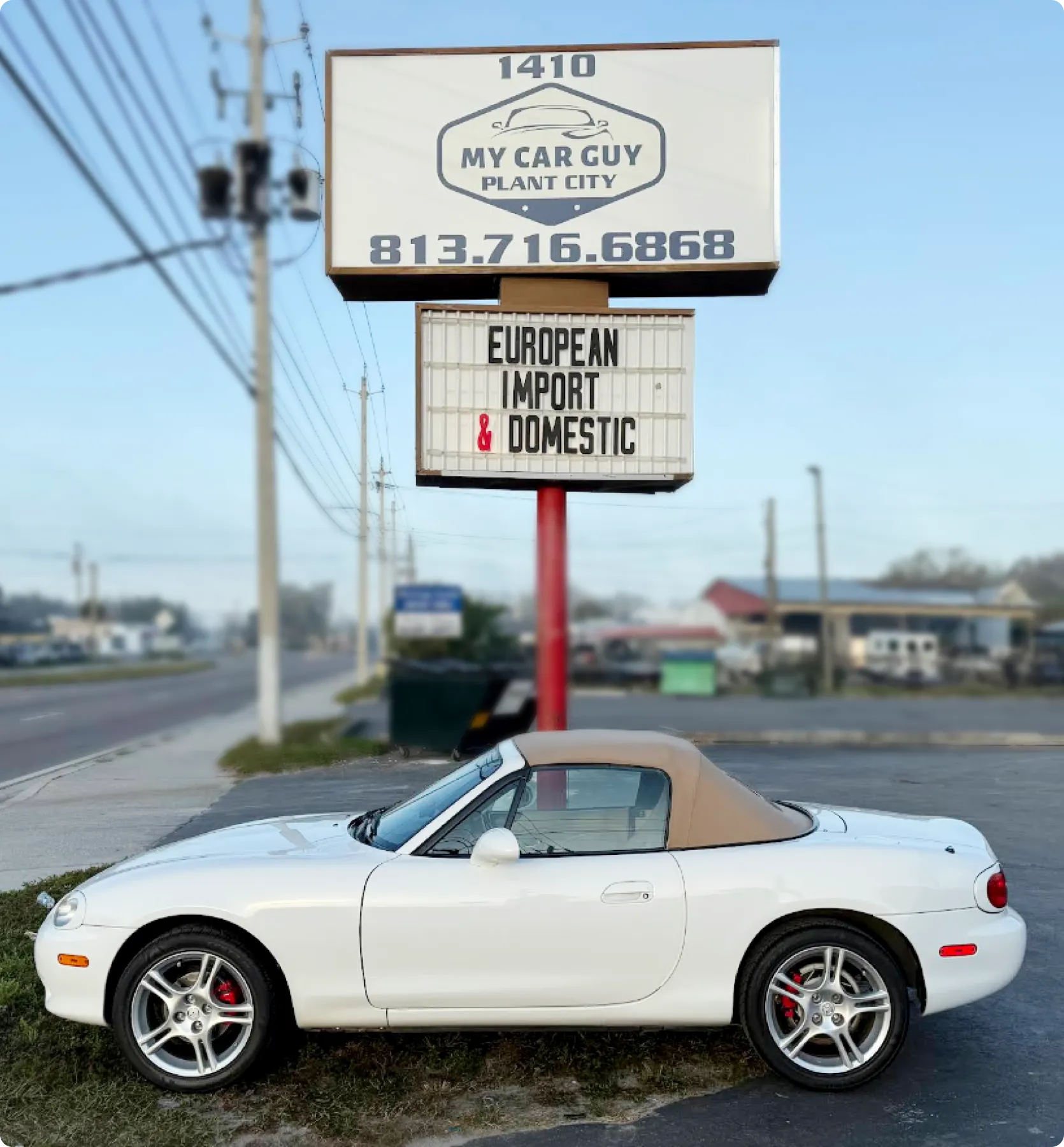 A white convertible car with a tan soft top is parked under a sign reading "My Car Guy Plant City" with contact info. Below, it says "European Import & Domestic."