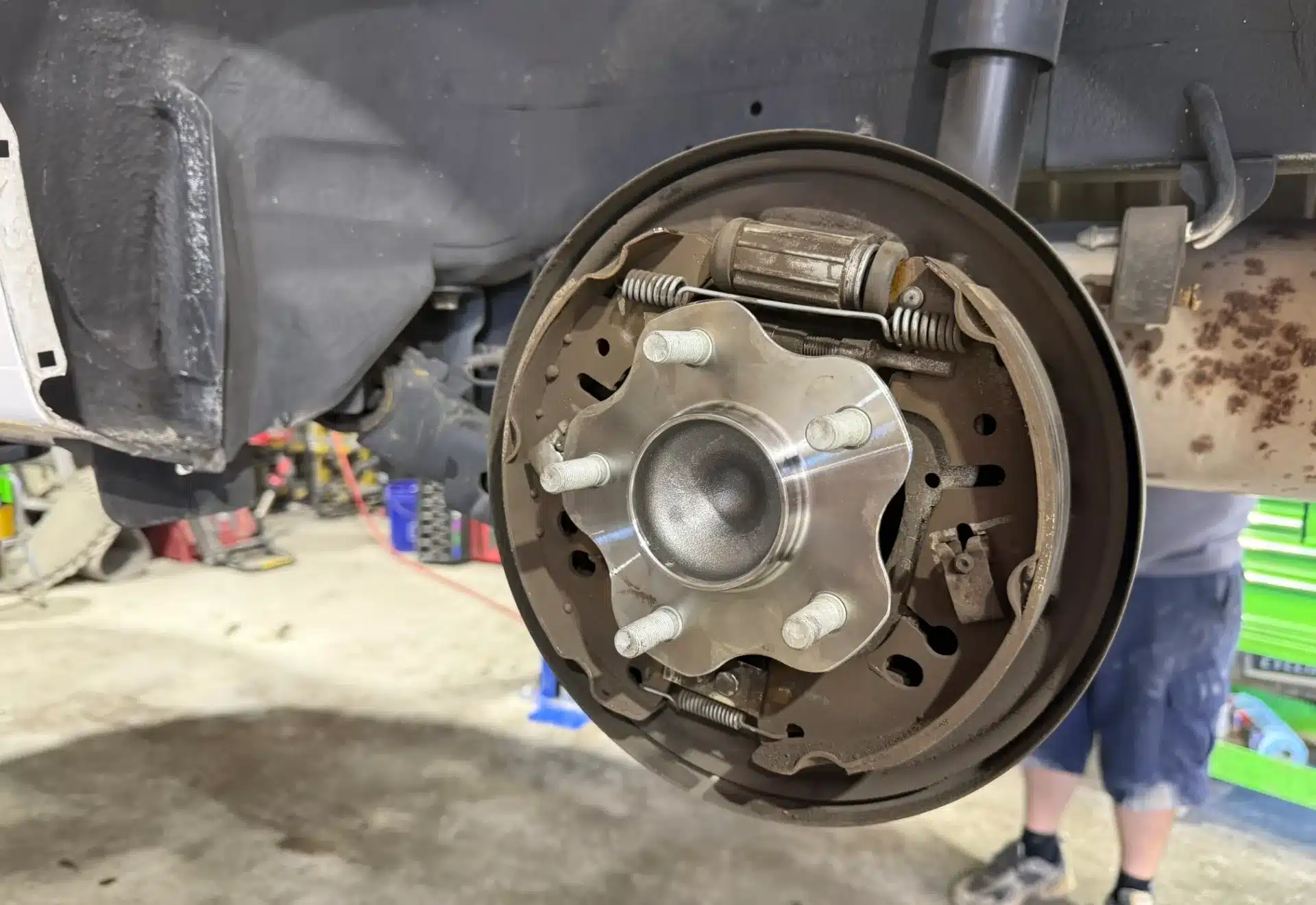 Close-up of a car's drum brake assembly with visible brake shoes, springs, and hub. A person in shorts and sneakers is blurred in the background.
