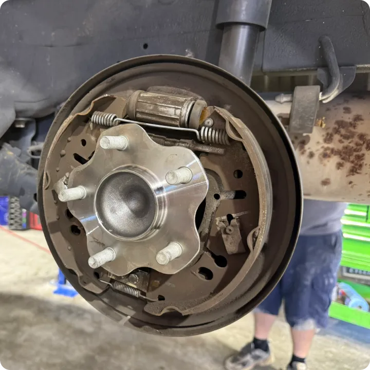 Close-up of a car's drum brake assembly with visible brake shoes, springs, and hub. A person in shorts and sneakers is blurred in the background.