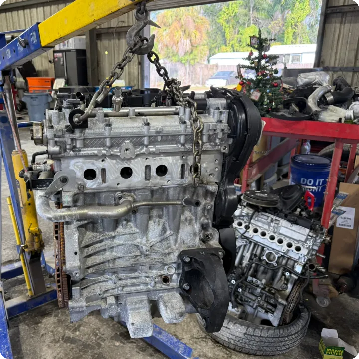 An engine is suspended on a hoist in a workshop, with tools and car parts scattered. A small decorated Christmas tree in the background adds a festive touch.