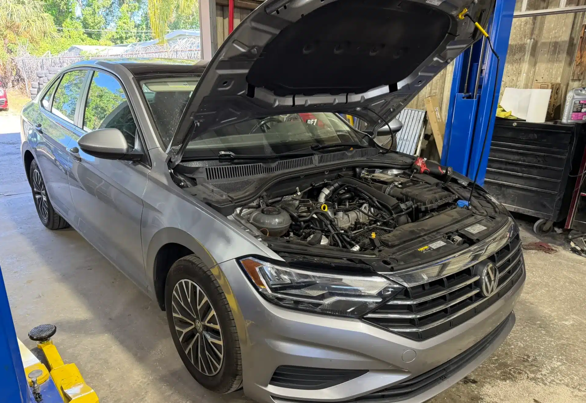 Silver Volkswagen car with the hood open, revealing the engine. The car is in a garage, conveying a setting of automotive repair or maintenance.