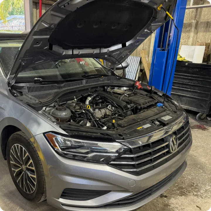 Silver Volkswagen car with the hood open, revealing the engine. The car is in a garage, conveying a setting of automotive repair or maintenance.