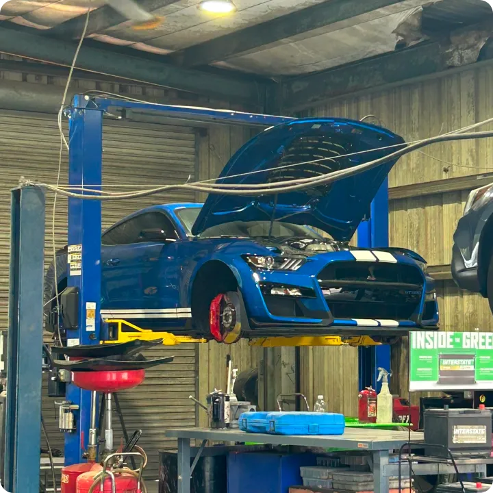 A blue car with its hood open is elevated on a hydraulic lift inside a garage, with a table full of tools below. The garage appears busy and industrial.
