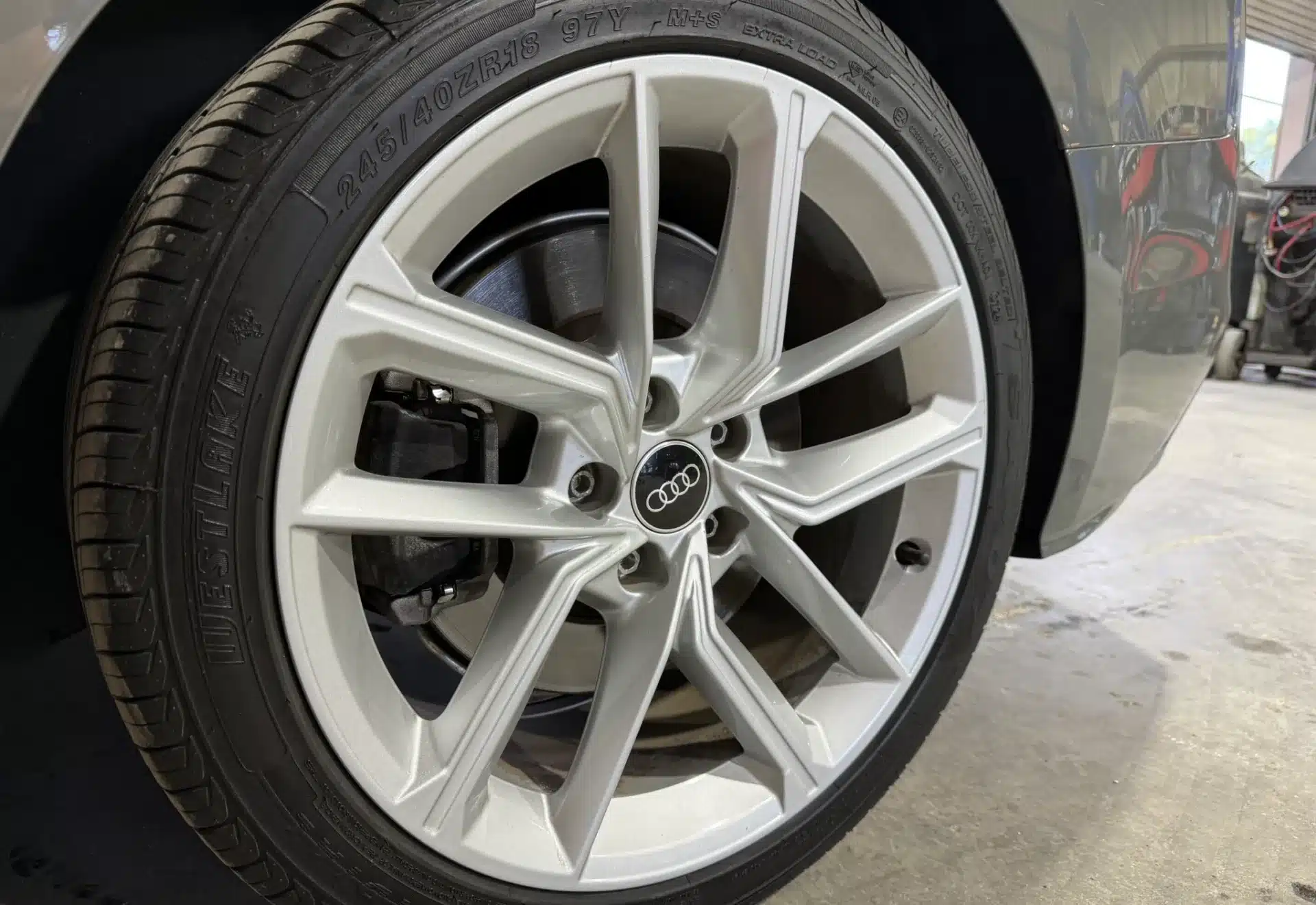 Close-up of a silver Audi car wheel showcasing the intricate, sleek alloy spokes and the brand logo at the center, parked on a concrete floor.