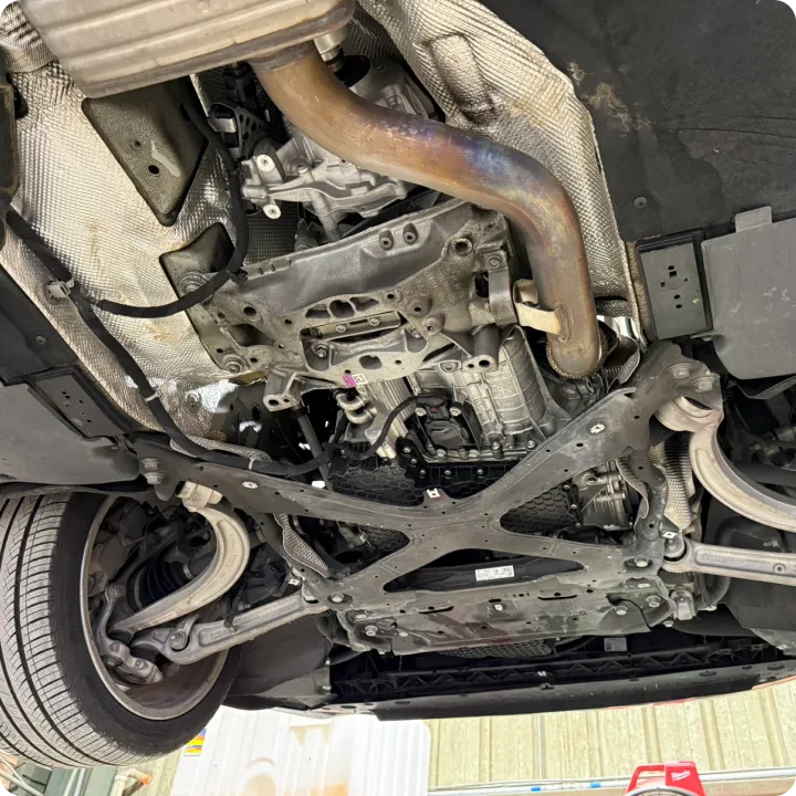 The image shows the underside of a vehicle on a lift, revealing the exhaust system, suspension, and parts of the chassis. The surroundings appear to be a garage.